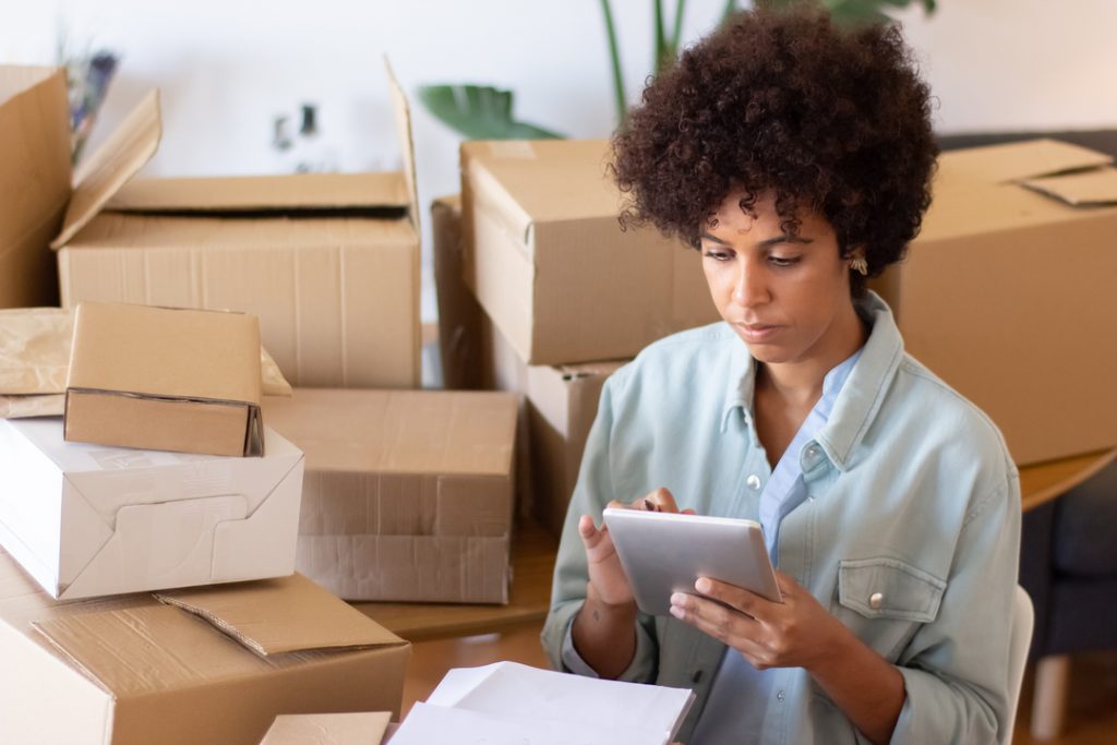 Gestão de entregas: imagem mostra mulher fazendo anotações, com caixas de papelão em volta.